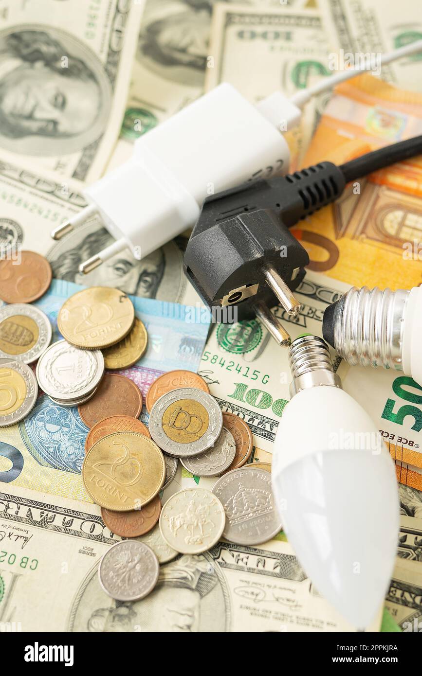 Il concetto di crisi energetica, i prezzi aumentano. Banconote in euro e in dollari, monete sullo sfondo di una lampada, spina, cavo. Tempo fiscale, pagamento fattura, calcolatore per il conteggio. Foto verticale. Foto Stock