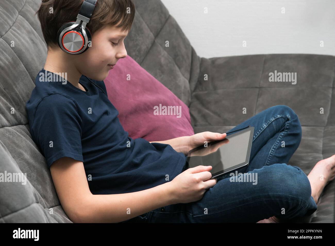 Happy boy con un tablet sul divano, vista laterale. I bambini in cuffia giocano a videogame su gadget o guardano un video. Foto Stock