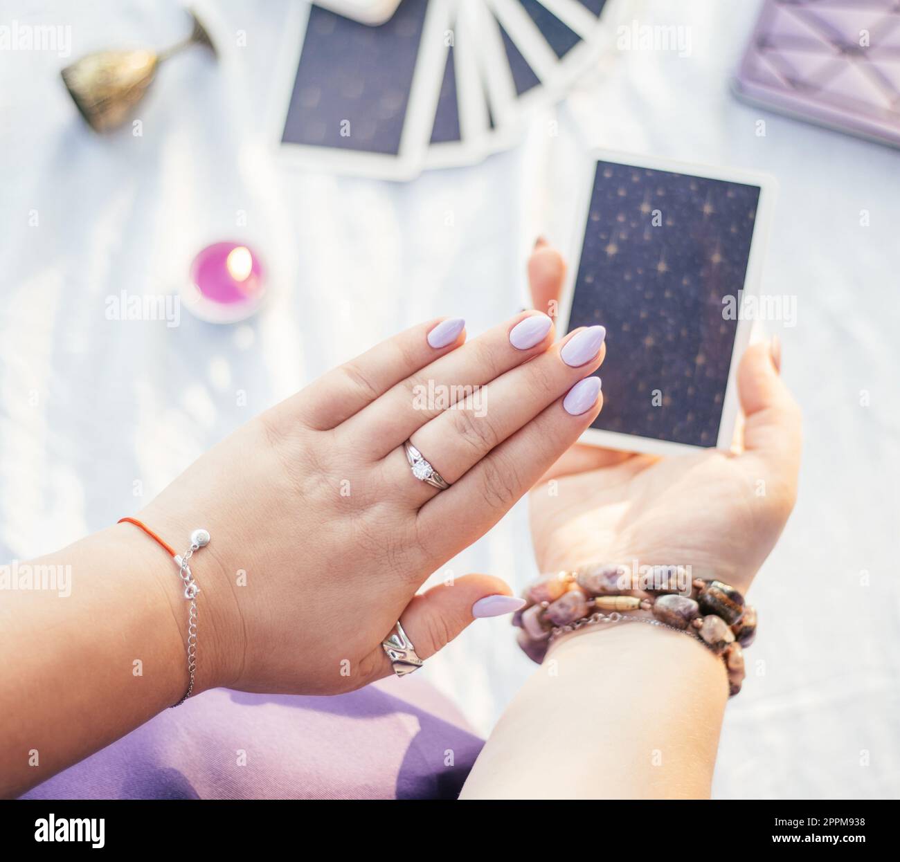 Donna tiene la mano con unghie viola su una carta Tarocchi su una superficie bianca con notebook e candela, vista dall'alto. Foto Stock