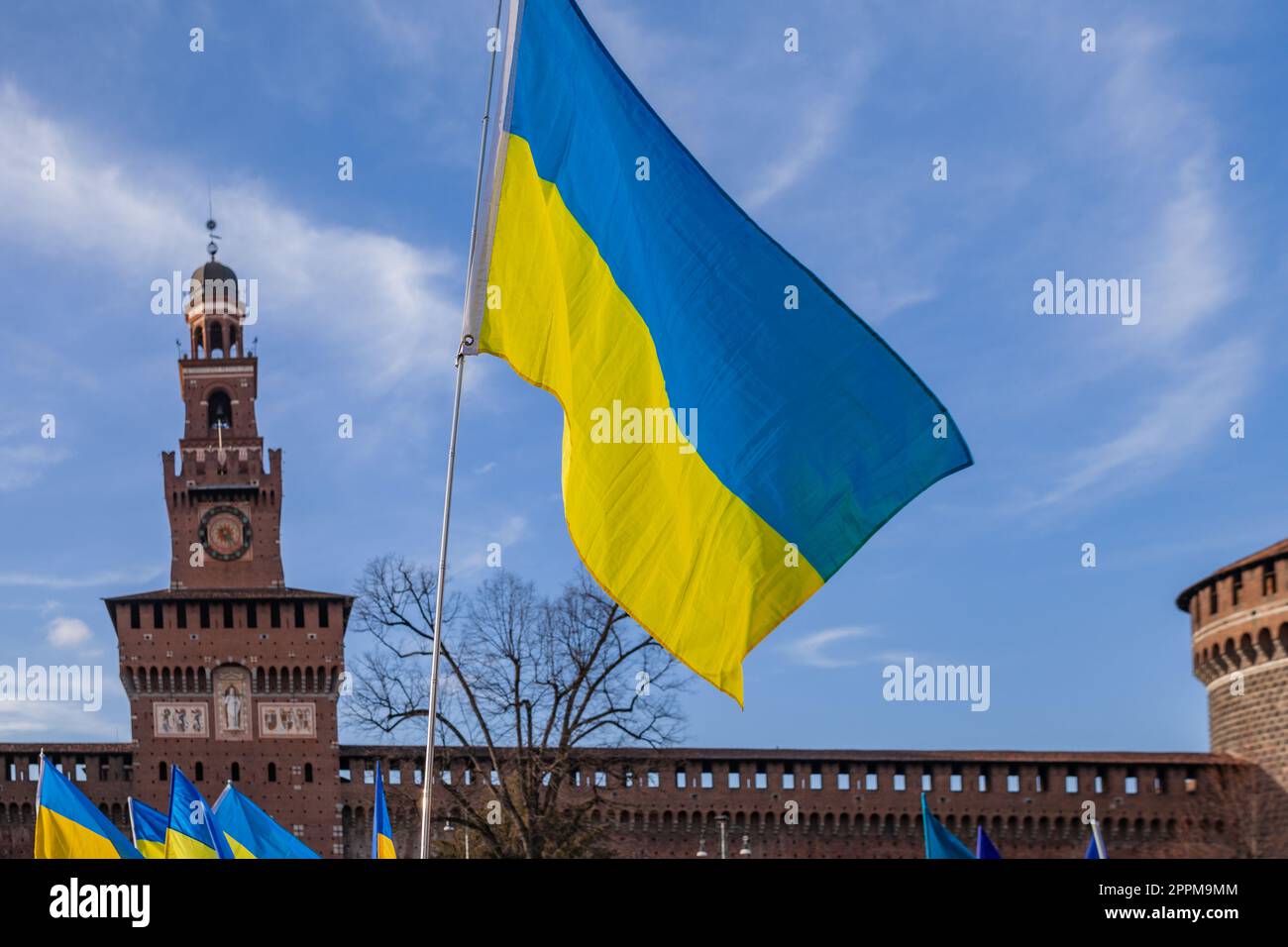 Bandiera con colori a strisce gialle e blu dell'Ucraina che ondola nel vento con un cielo e un sole blu. Sullo sfondo il Castello Sforzesco di Milano. Foto Stock