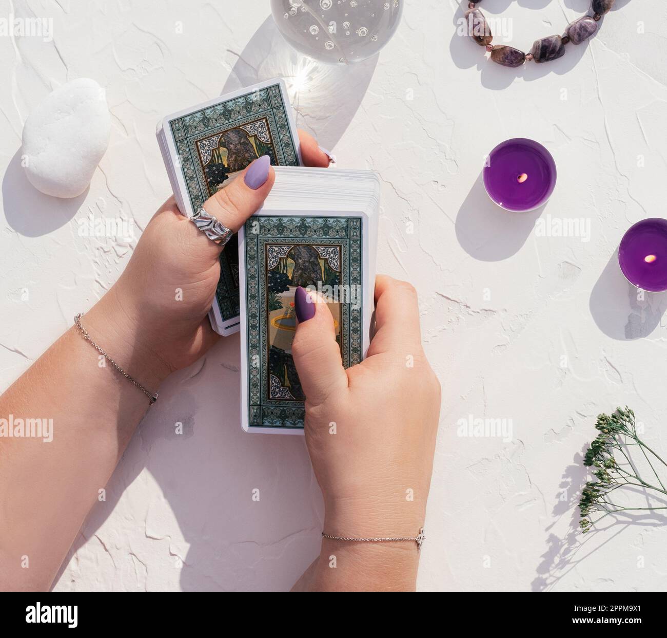 Le mani con chiodi e anelli viola reggono un mazzo di carte Tarocchi su una superficie bianca. Sfera di cristallo, candele e pietre, vista dall'alto Foto Stock