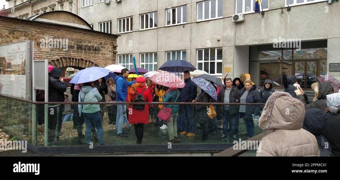 Sarajevo, Bosnia ed Erzegovina, 8 marzo 2020. Un gruppo di turisti che ascoltano la guida. Piove, la gente sta sotto vari ombrelli. Antichi edifici in pietra. Bezistan Foto Stock
