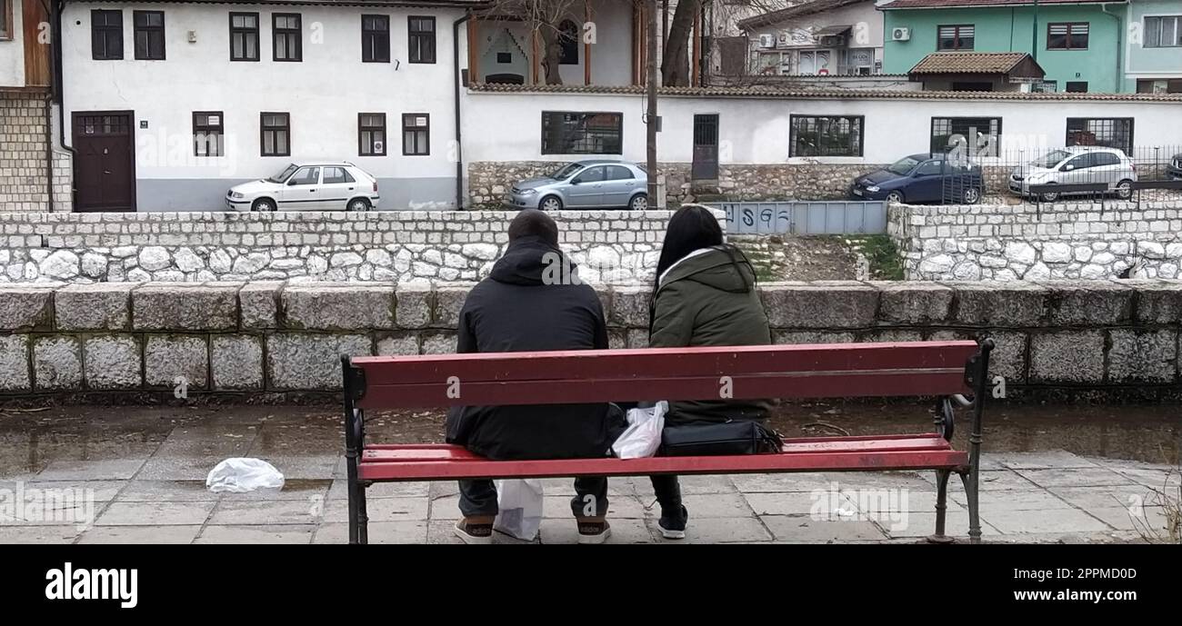 Sarajevo, Bosnia ed Erzegovina - 08 marzo 2020. Una giovane coppia seduta su una panchina di legno sotto un albero. L'argine del fiume Milyack. Vista sulla città di Sarajevo, una moschea e un minareto. Foto Stock