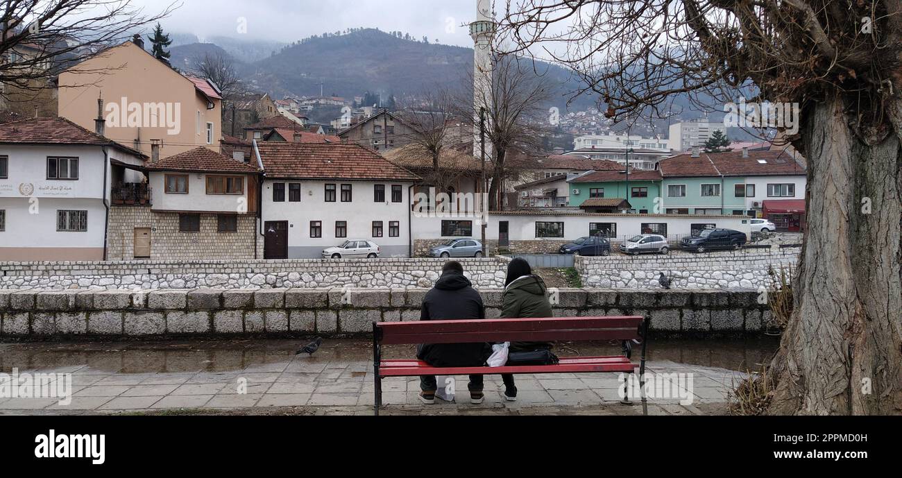 Sarajevo, Bosnia ed Erzegovina - 08 marzo 2020. Una giovane coppia seduta su una panchina di legno sotto un albero. L'argine del fiume Milyack. Vista sulla città di Sarajevo, una moschea e un minareto Foto Stock