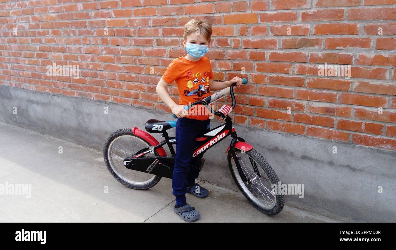 Belgrado, Serbia, 13 aprile 2020: Un bel ragazzo biondo di 7 anni indossa una maschera chirurgica protettiva blu sul suo viso. Il bambino va in bicicletta, fa una sosta presso un muro di mattoni e guarda la macchina fotografica Foto Stock