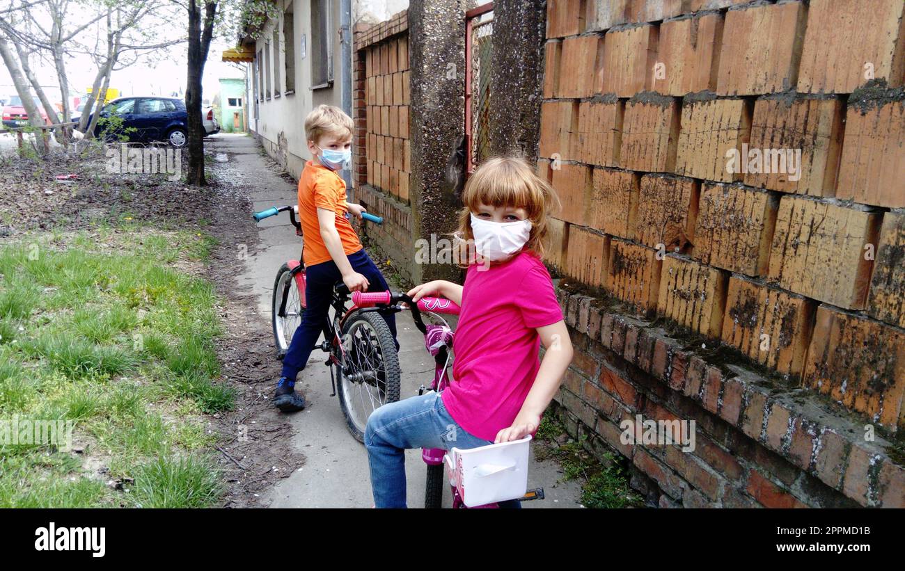 I bambini di 6 e 7 anni con maschere chirurgiche protettive bianche entrano per andare in bicicletta. Mettere in pausa il ciclo. Una ragazza con una maglietta rosa e un ragazzo biondo in arancione. Bambini per strada vicino alla vecchia recinzione in mattoni Foto Stock