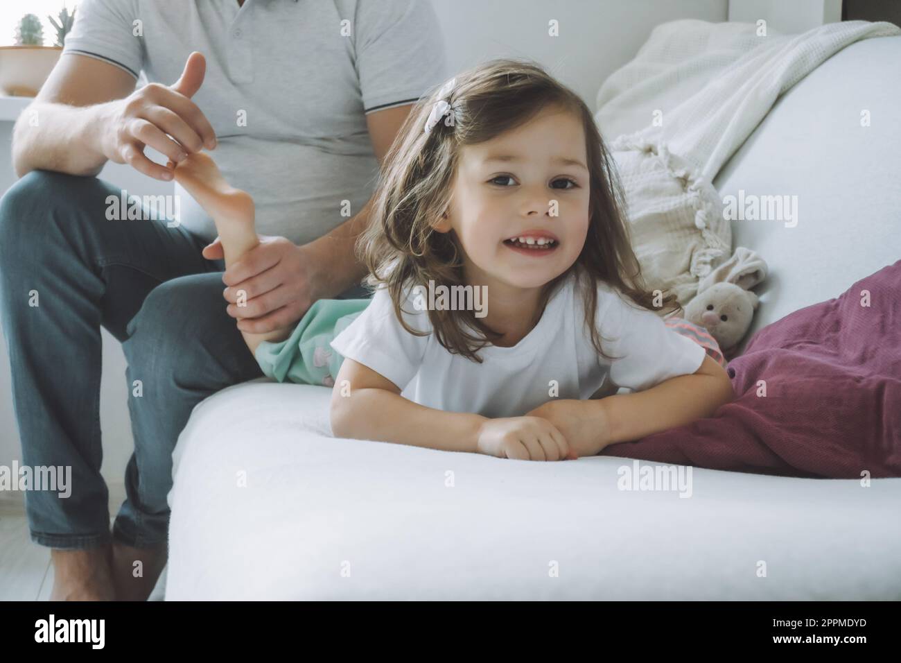 Il padre gioca con la figlia piccola 2-4 sul divano. Papà fa il solletico ai piedi dei bambini. Famiglia, divertimento Foto Stock