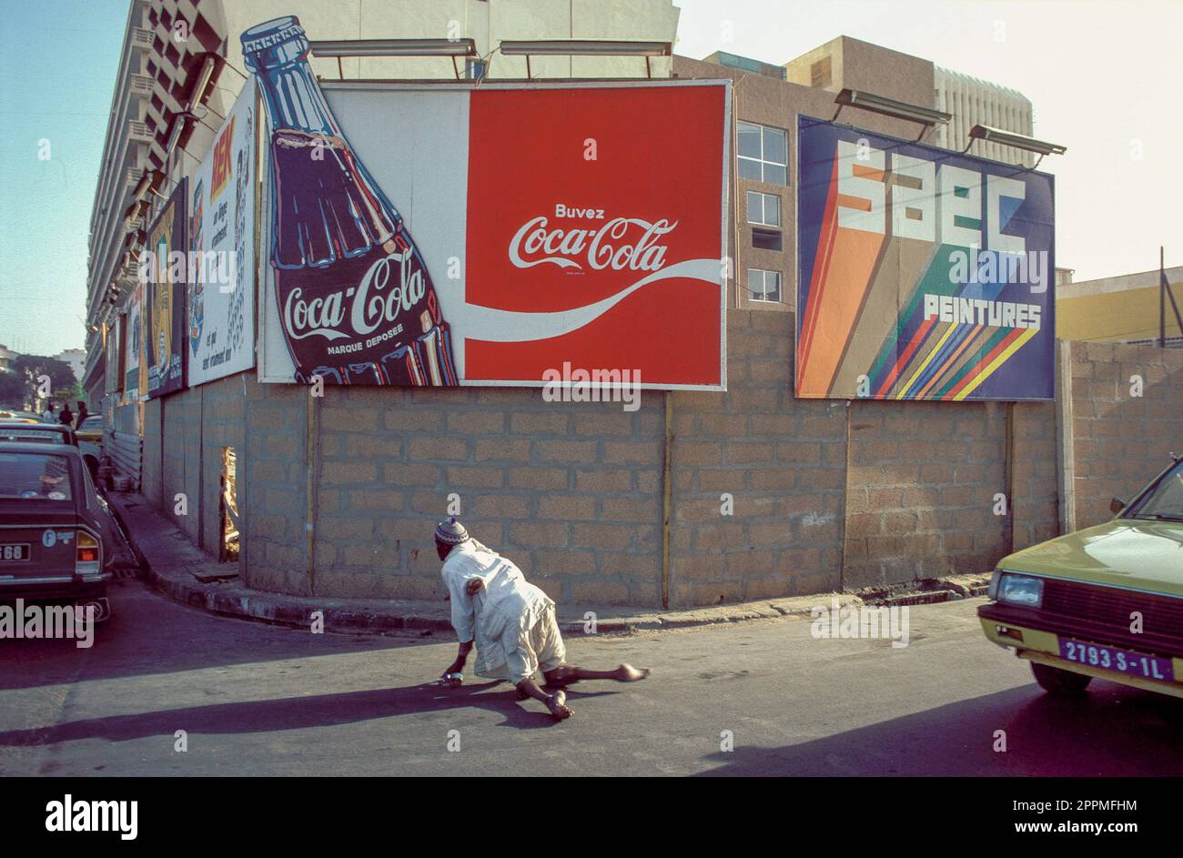 Senegal, Un contrasto tra il cartellone della coca cola e un mendicante che sta strisciando sulla strada di fronte ad esso. Foto Stock