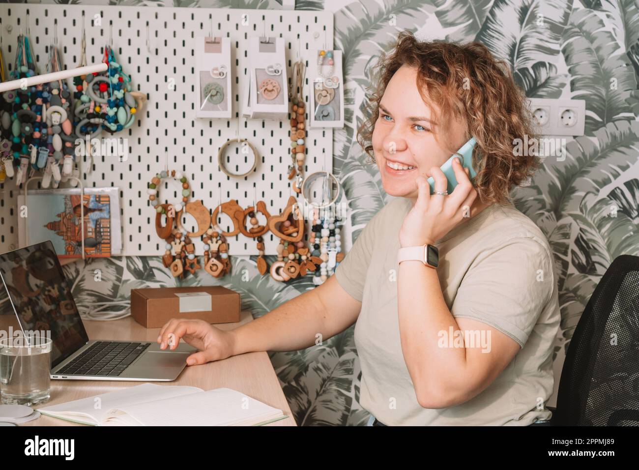 Sorridente donna d'affari concentrata che parla di smartphone, che lavora su un computer portatile in officina. Ufficio domestico. Accessori per bambini Foto Stock