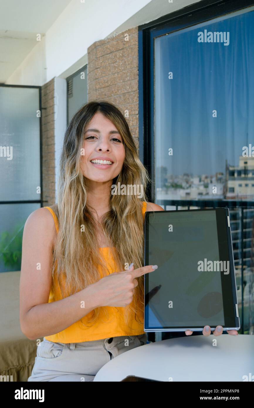 immagine verticale di una giovane donna d'affari latina, di etnia colombiana, sorridente seduto guardando la macchina fotografica che indica lo schermo del portatile e guardando il camer Foto Stock