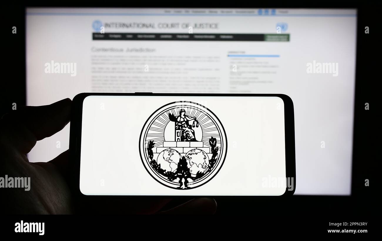 Persona che tiene il cellulare con il logo della Corte Internazionale di Giustizia (ICJ) sullo schermo di fronte alla pagina web. Messa a fuoco sul display del telefono. Foto Stock