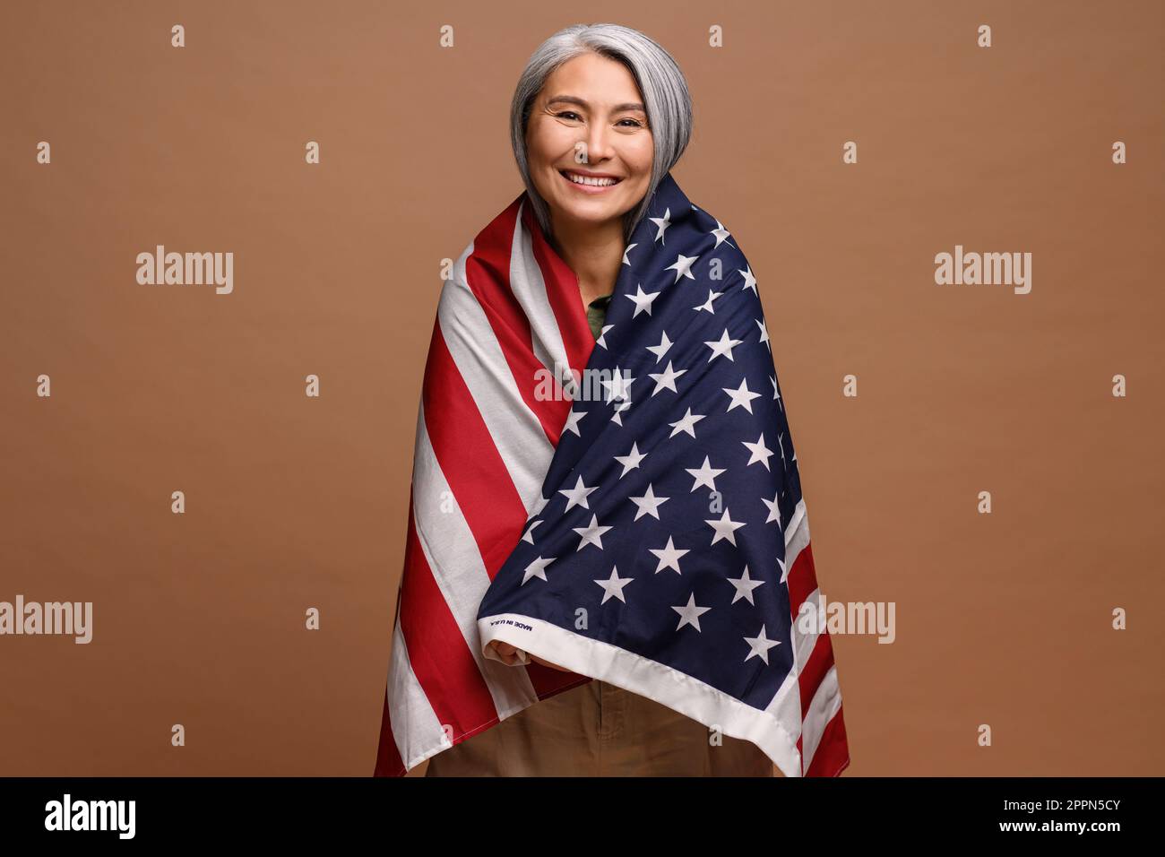 Allegro grigio-capelli 50s donna asiatica che avvolge la bandiera degli Stati Uniti isolato su sfondo marrone. Grande donna patriottica celebrata il 4th luglio Foto Stock