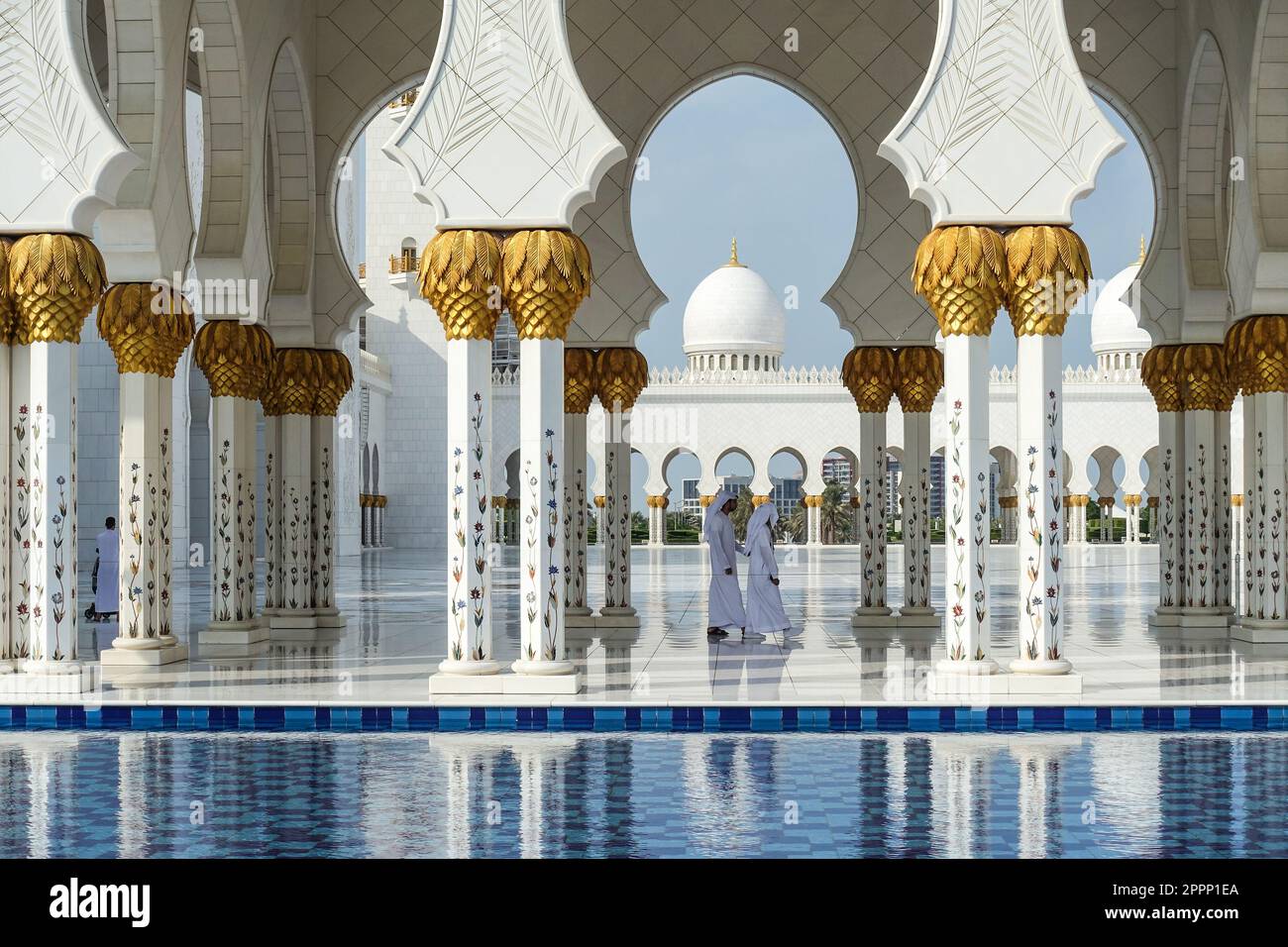 I visitatori camminano attraverso il colonnato ornato della grande Moschea Sheikh Zayed caratterizzato da marmo bianco, accenti d'oro, archi e cupole riflesse nell'acqua Foto Stock