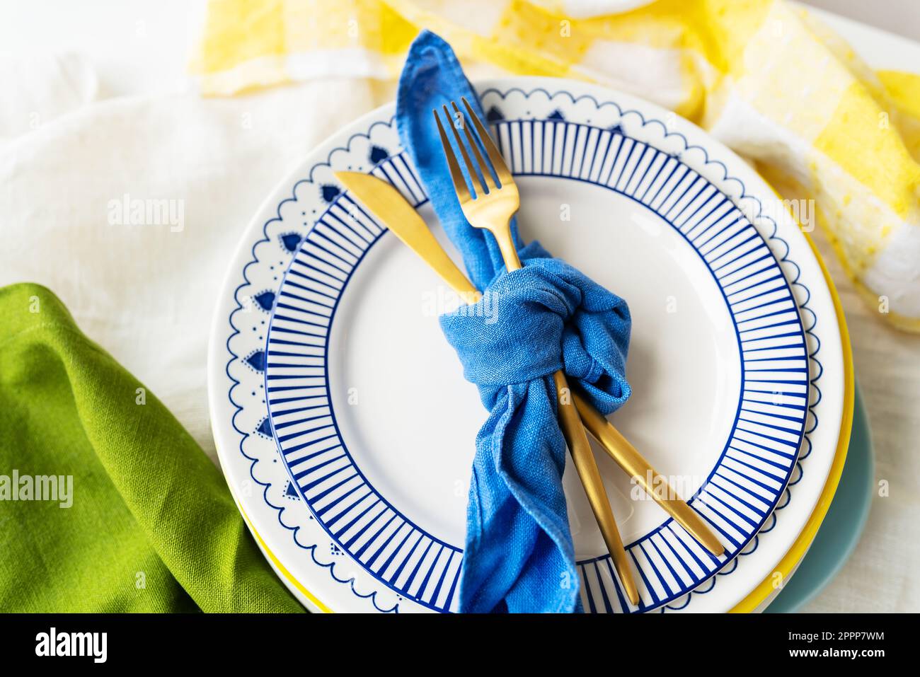 Design di servizio. Concetto di servizio di Pasqua. Le posate dorate sono legate con un tovagliolo blu, su cui i piatti sono su un tovagliolo di lino verde-giallo. Minimalista Foto Stock