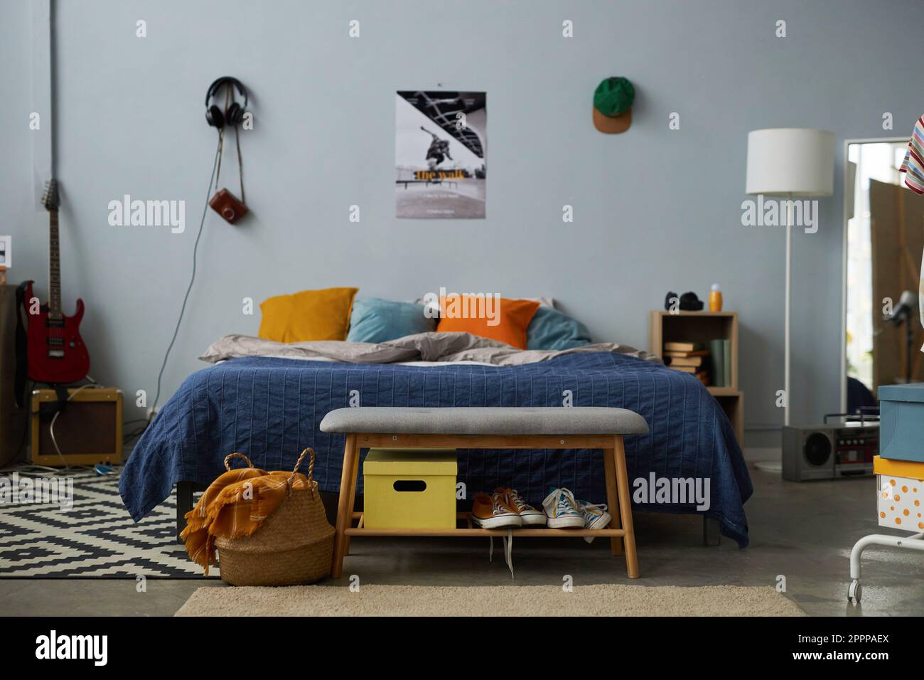 Vista frontale di un comodo letto matrimoniale con cuscini, lampada e chitarra in piedi contro la parete grigia con poster, cuffie e cuffia Foto Stock