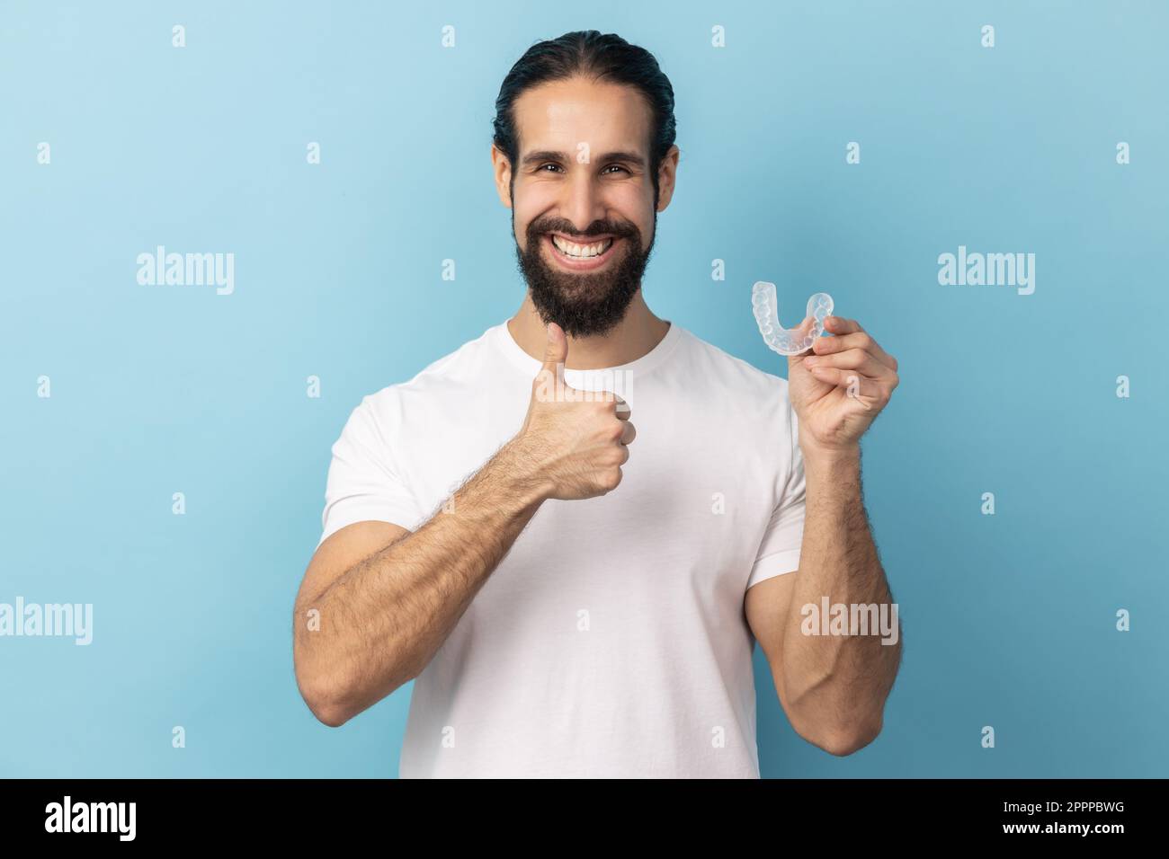 Ritratto dell'uomo con barba che indossa una T-shirt bianca che tiene il fermo dell'allineatore dentale, clinica dentale per i bei denti, mostrando il pollice in alto. Studio in interni isolato su sfondo blu. Foto Stock