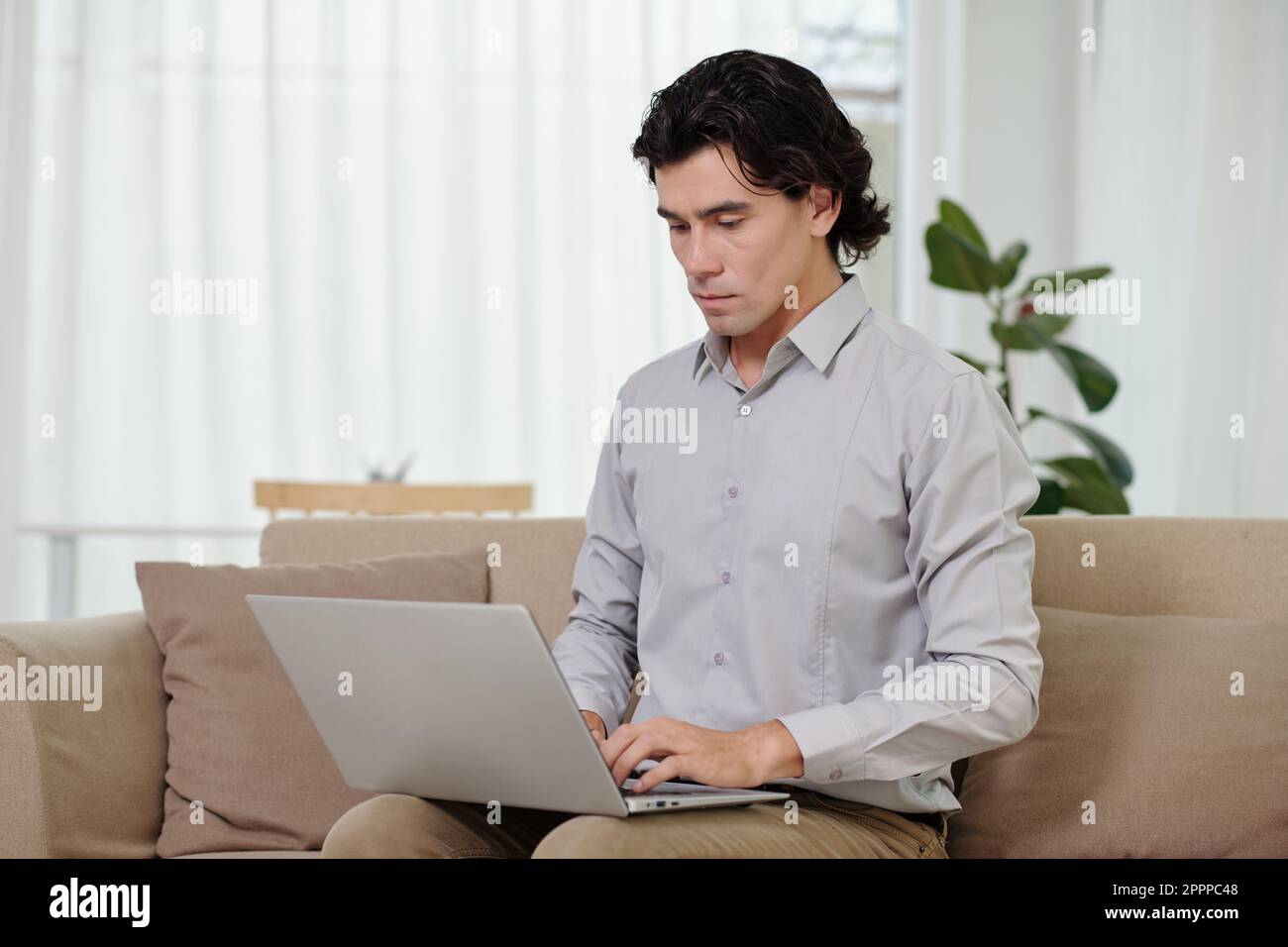 Analista giovane maschio che digita sulla tastiera del notebook mentre si siede sul divano in ufficio a casa e comunica con un collega online Foto Stock