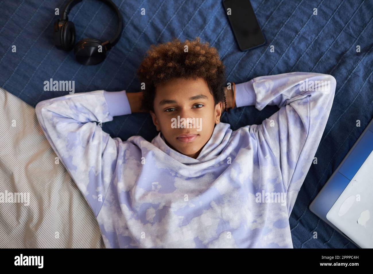 Ragazzo giovane e sereno in elegante felpa con cappuccio che guarda la macchina fotografica mentre sdraiato sul morbido letto matrimoniale blu e rilassante dopo aver fatto i compiti a casa Foto Stock