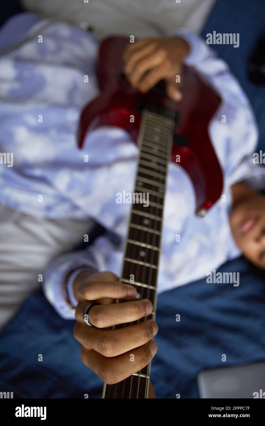 Concentratevi sulla mano di un giovane musicista maschio su corde di chitarra elettrica suonandola mentre vi godete il piacere di un comodo letto a casa Foto Stock