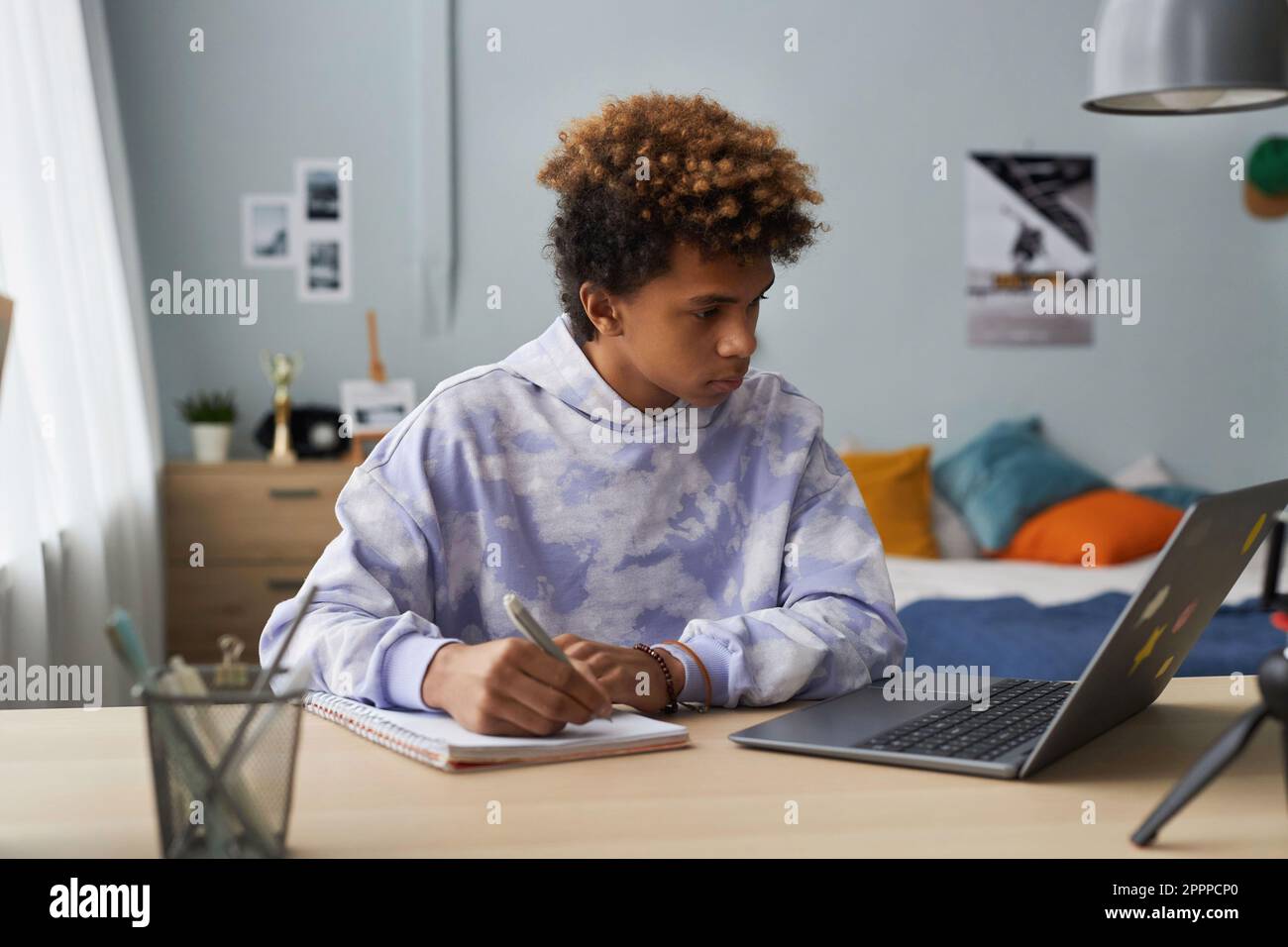 Studente serio adolescente che prende appunti in copybook mentre si siede alla scrivania di fronte al computer portatile e l'ascolto di techer durante la lezione online Foto Stock