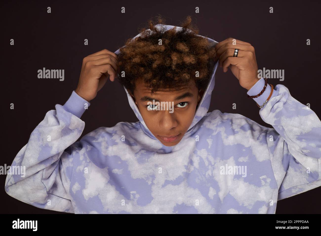 Ragazzo afro-americano giovane mettere su cappuccio sulla sua testa e guardare la macchina fotografica mentre si trova su sfondo nero in isolamento in studio Foto Stock