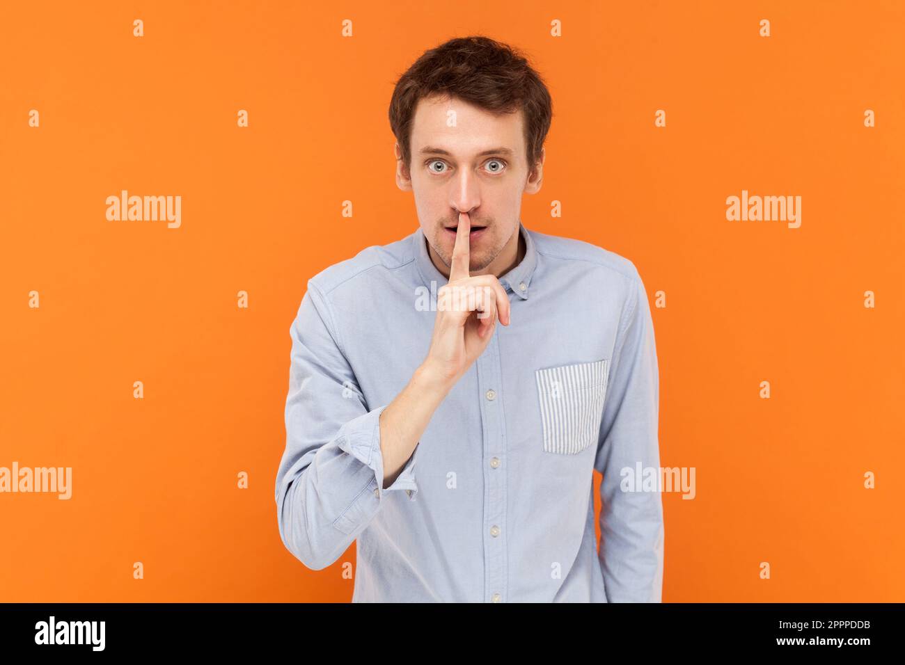 Serio giovane uomo adulto in piedi con il dito vicino alle labbra, chiedendo di tenersi in silenzio, guardando la macchina fotografica con espressione bossy, indossando una camicia blu chiaro. Studio al coperto isolato su sfondo arancione. Foto Stock