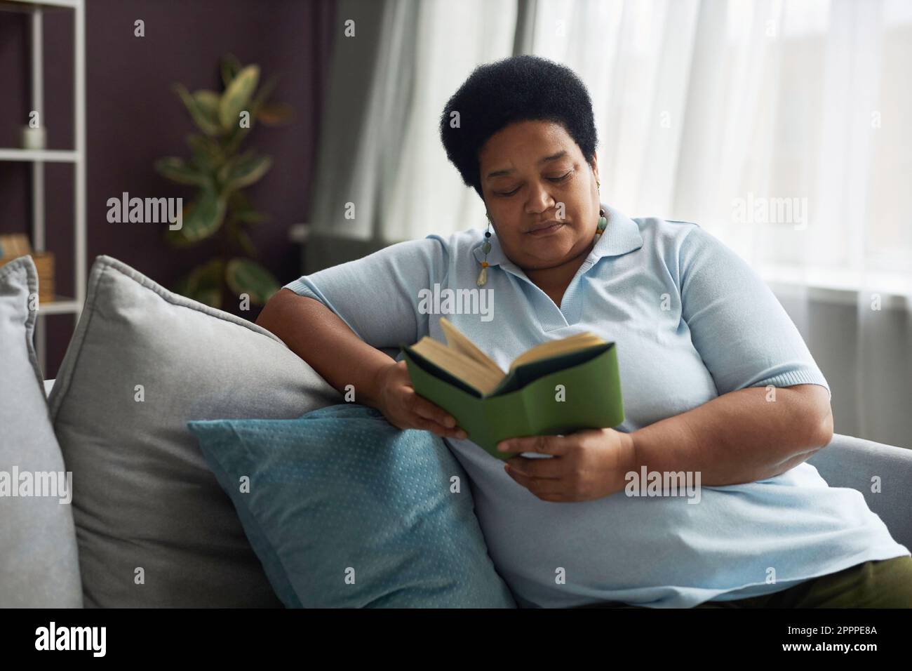 Donna afro-americana matura più grande in casualwear seduta sul divano con cuscini e libro di lettura mentre si soggiorna a casa il fine settimana Foto Stock