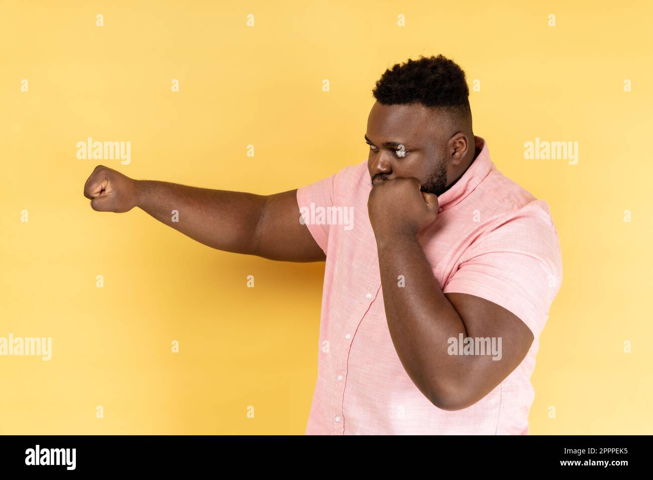 Ti colpisco. Vista laterale dell'uomo bearded che indossa una camicia rosa in piedi con pugilato in rilievo pronto a pugni, pronto a combattere, spirito di combattimento. Studio al coperto isolato su sfondo giallo Foto Stock