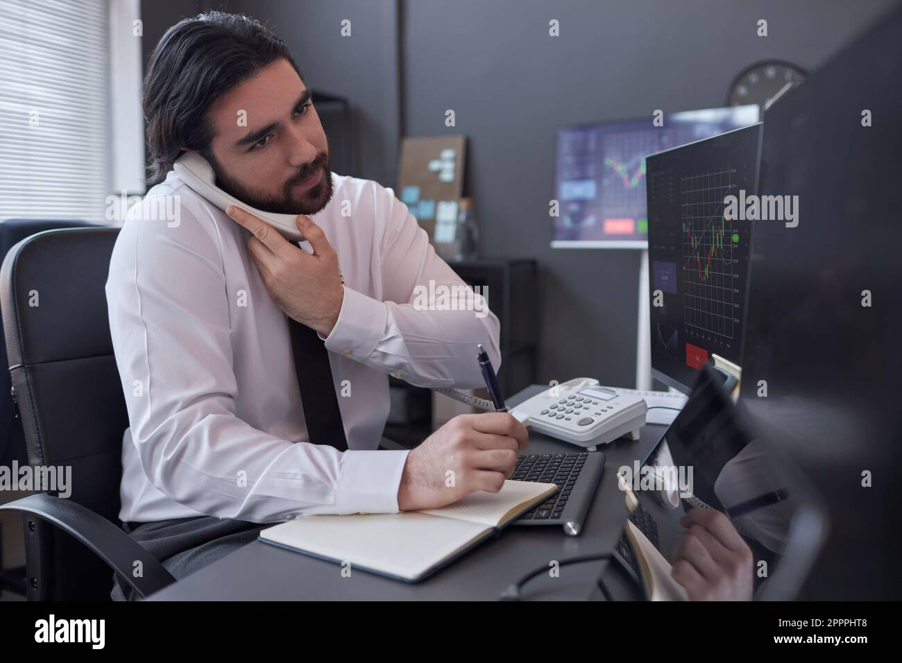 Un giovane broker che si rivolge al cliente per consultare la situazione del mercato azionario al telefono seduto sul suo posto di lavoro con computer Foto Stock
