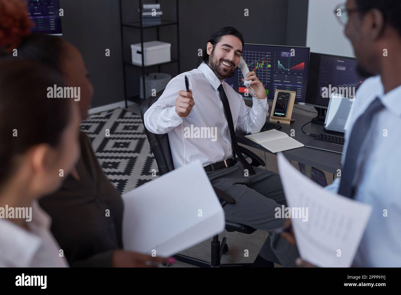 Giovane broker sorridente che vende le scorte con successo al cliente durante la conversazione al telefono Foto Stock