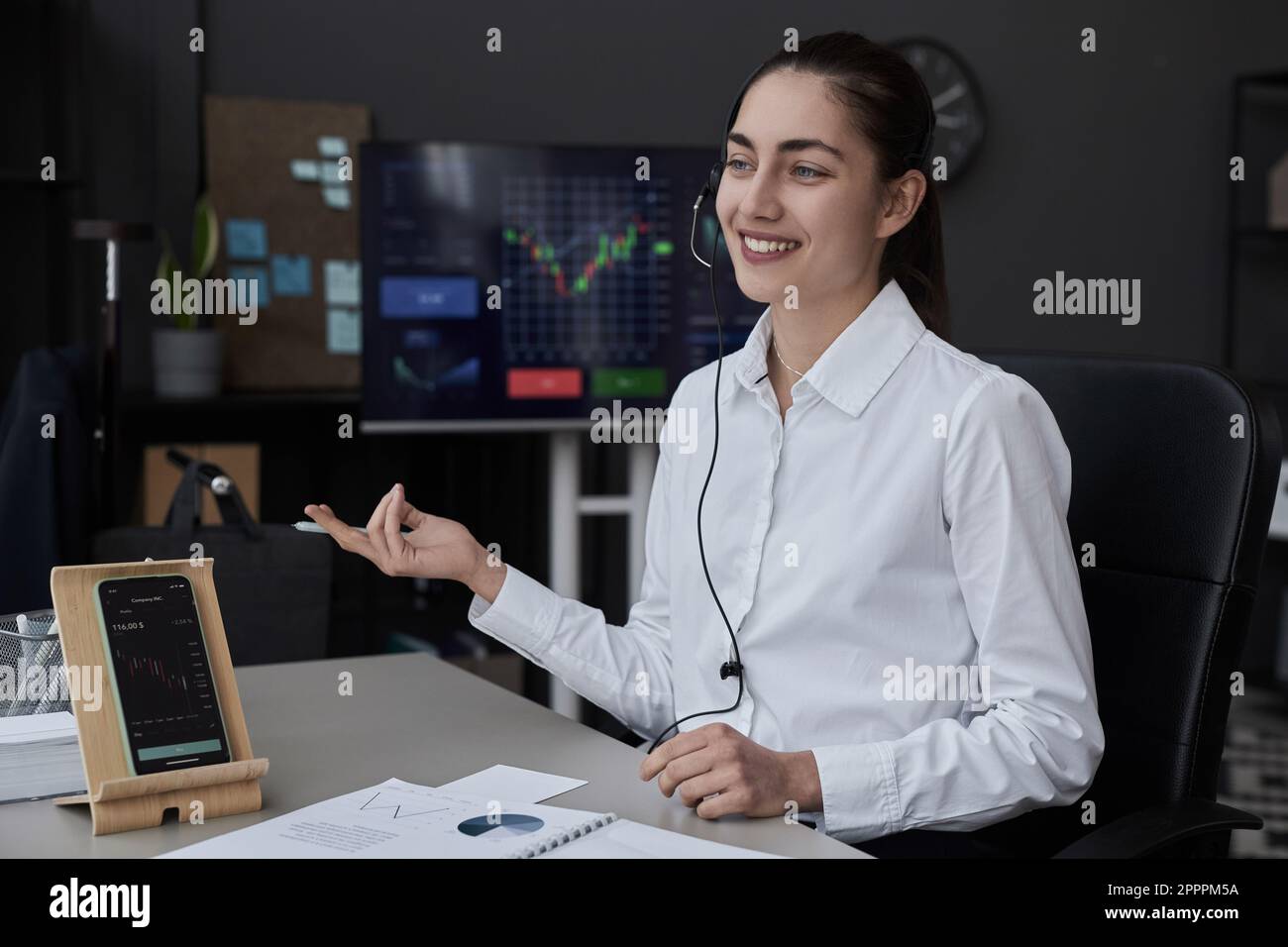 Giovane commerciante sorridente in cuffie che parlano con il cliente e discutono della situazione finanziaria in ufficio Foto Stock