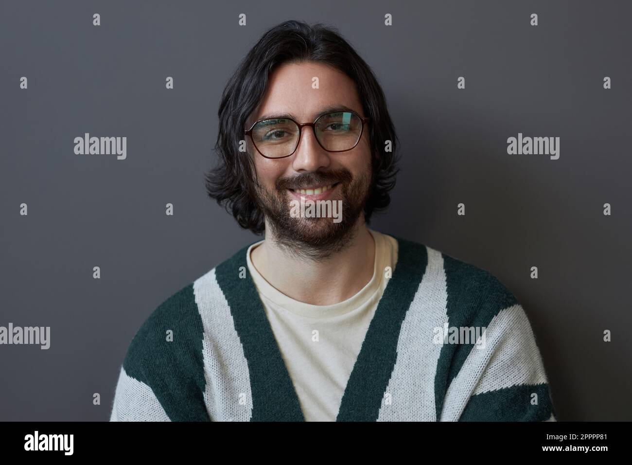 Ritratto di bell'uomo sorridente in occhiali con capelli lunghi sorridenti alla macchina fotografica su sfondo grigio Foto Stock