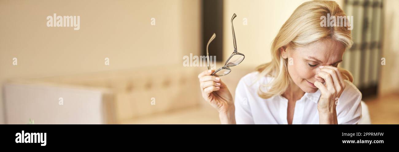 Donna d'affari stanca matura tolse i suoi occhiali, massaggiando il ponte nasale, soffrendo di sindrome degli occhi secchi Foto Stock