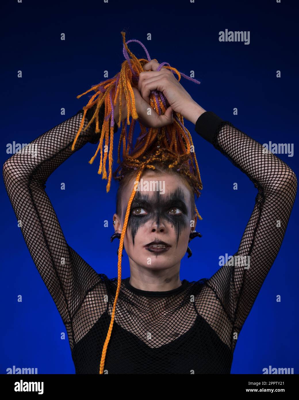 Informale giovane donna con capelli a treccia colorati e trucco palco nero spettrale dipinto sul viso. Vista frontale, ripresa da studio su sfondo blu. Parte del ph Foto Stock