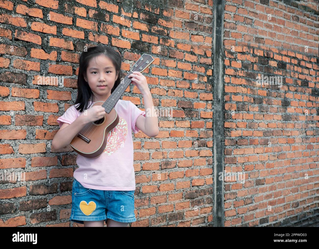 Piccolo bambino asiatico giocare l'ukulele, vecchio sfondo muro di mattoni Foto Stock