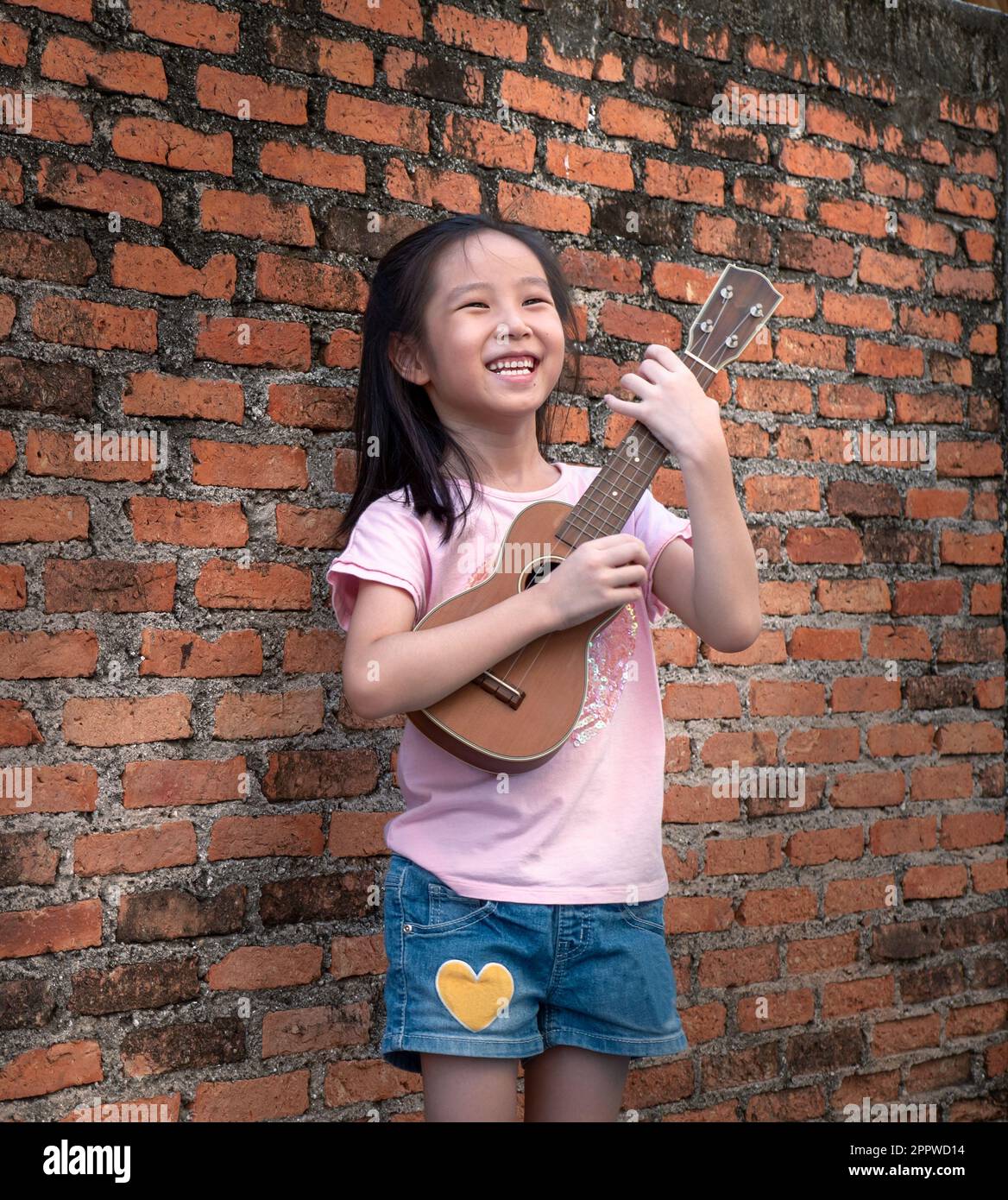 Piccolo bambino asiatico giocare l'ukulele, vecchio sfondo muro di mattoni Foto Stock