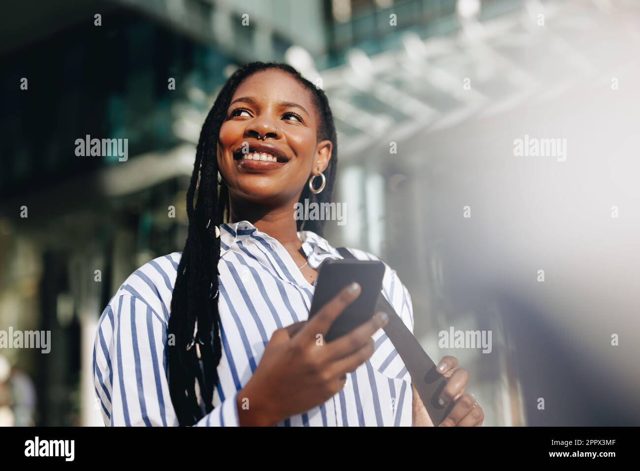Giovane donna d'affari pensosa che guarda via con un sorriso mentre si va a lavorare in città. Felice giovane donna d'affari che usa uno smartphone durante il suo viaggio t Foto Stock