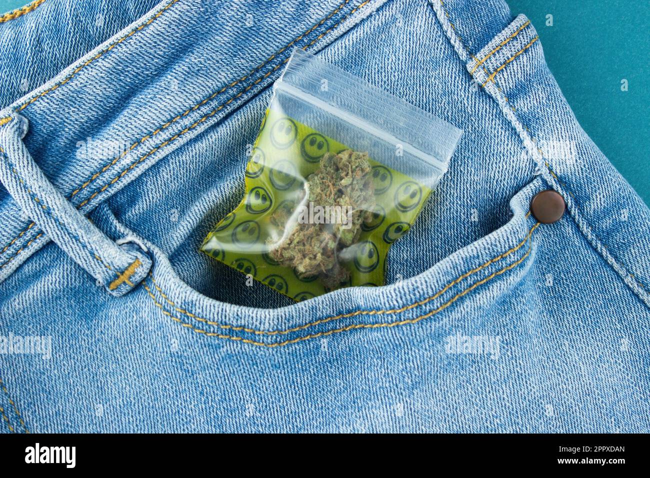 Un bocciolo di marijuana secco in un emoji con chiusura lampo fuoriesce dalla tasca anteriore dei jeans blu. Primo piano Foto Stock