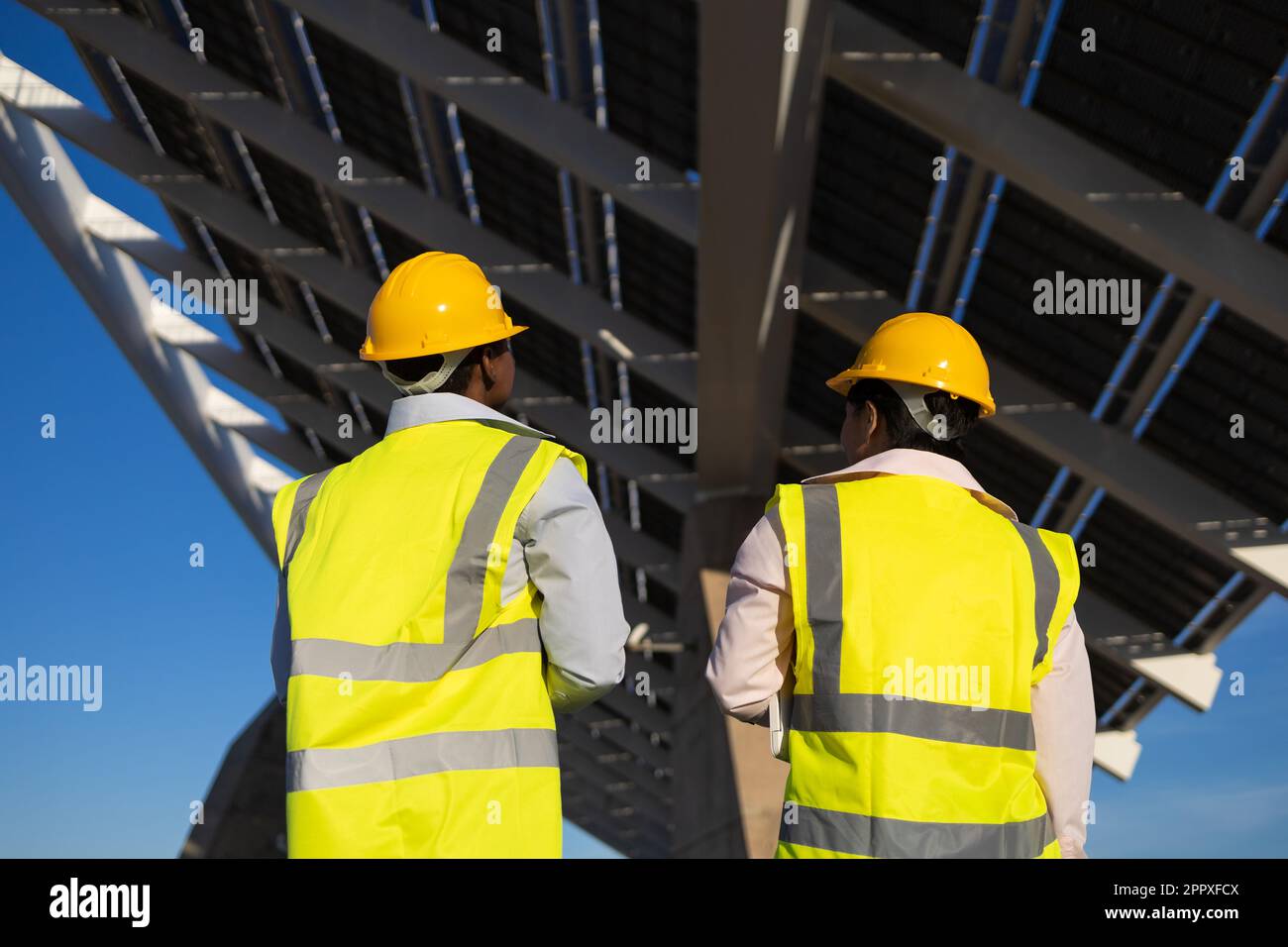 Vista posteriore di ingegneri anonimi in hardhat e uniforme in piedi vicino a pannelli solari installati in cantiere Foto Stock