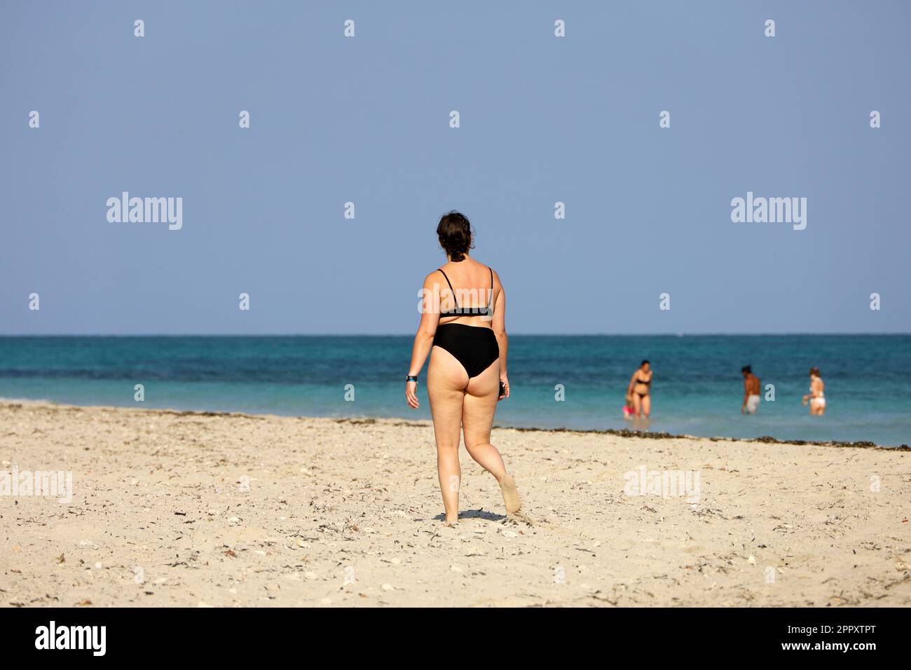 Donna in costume da bagno nero che cammina sulla sabbia sullo sfondo del mare. Vacanza in spiaggia, concetto di sovrappeso e di dieta Foto Stock