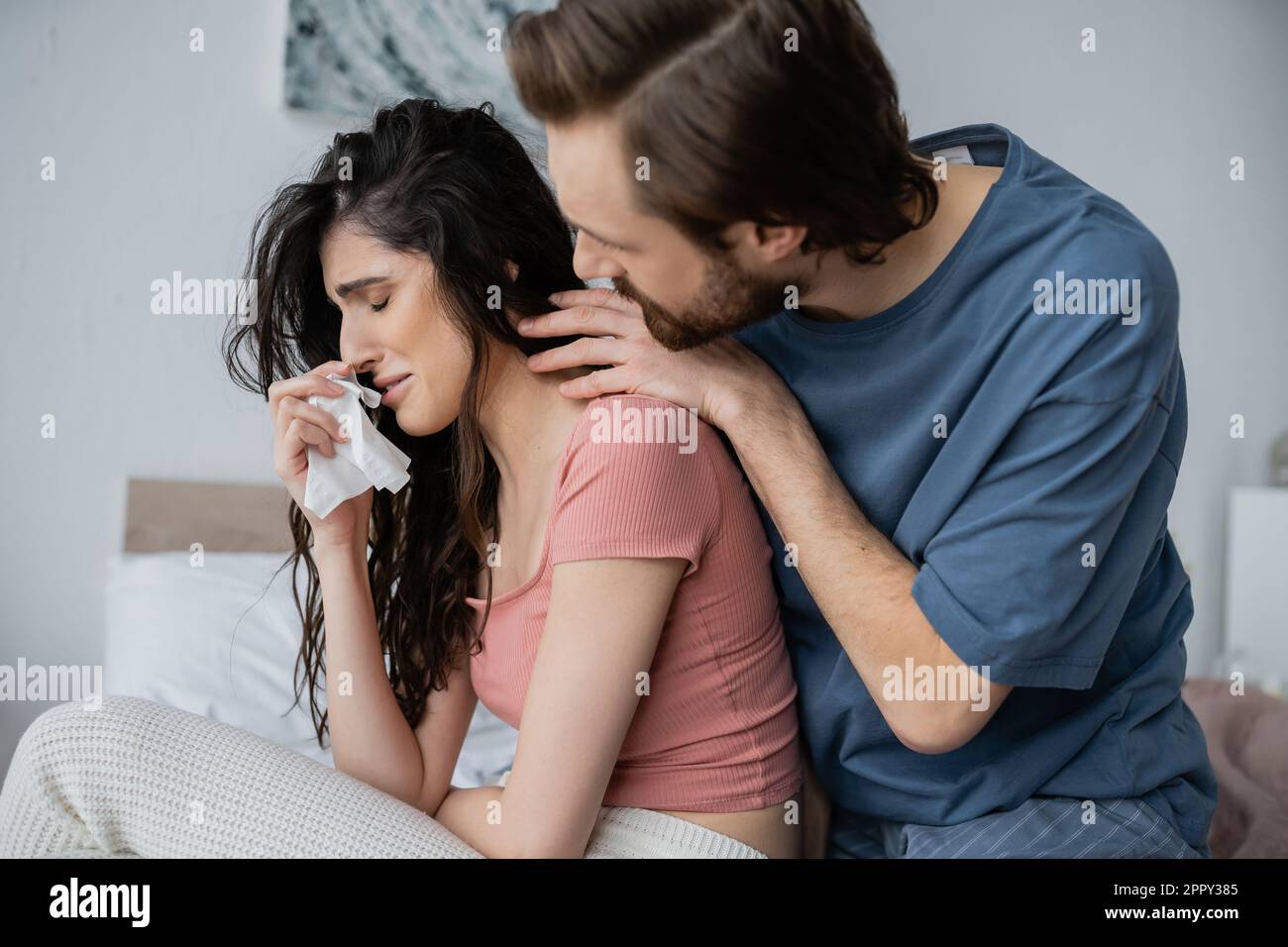Uomo attento in pajama abbracciando ragazza piangente con tovagliolo in camera da letto, immagine stock Foto Stock