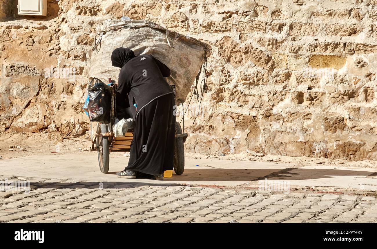 Sousse, Tunisia, 18 gennaio 2023: Donna anziana in abiti neri tradizionali con abaya e hijab spinge un carrello su cui si trova un sacco con raccolto Foto Stock