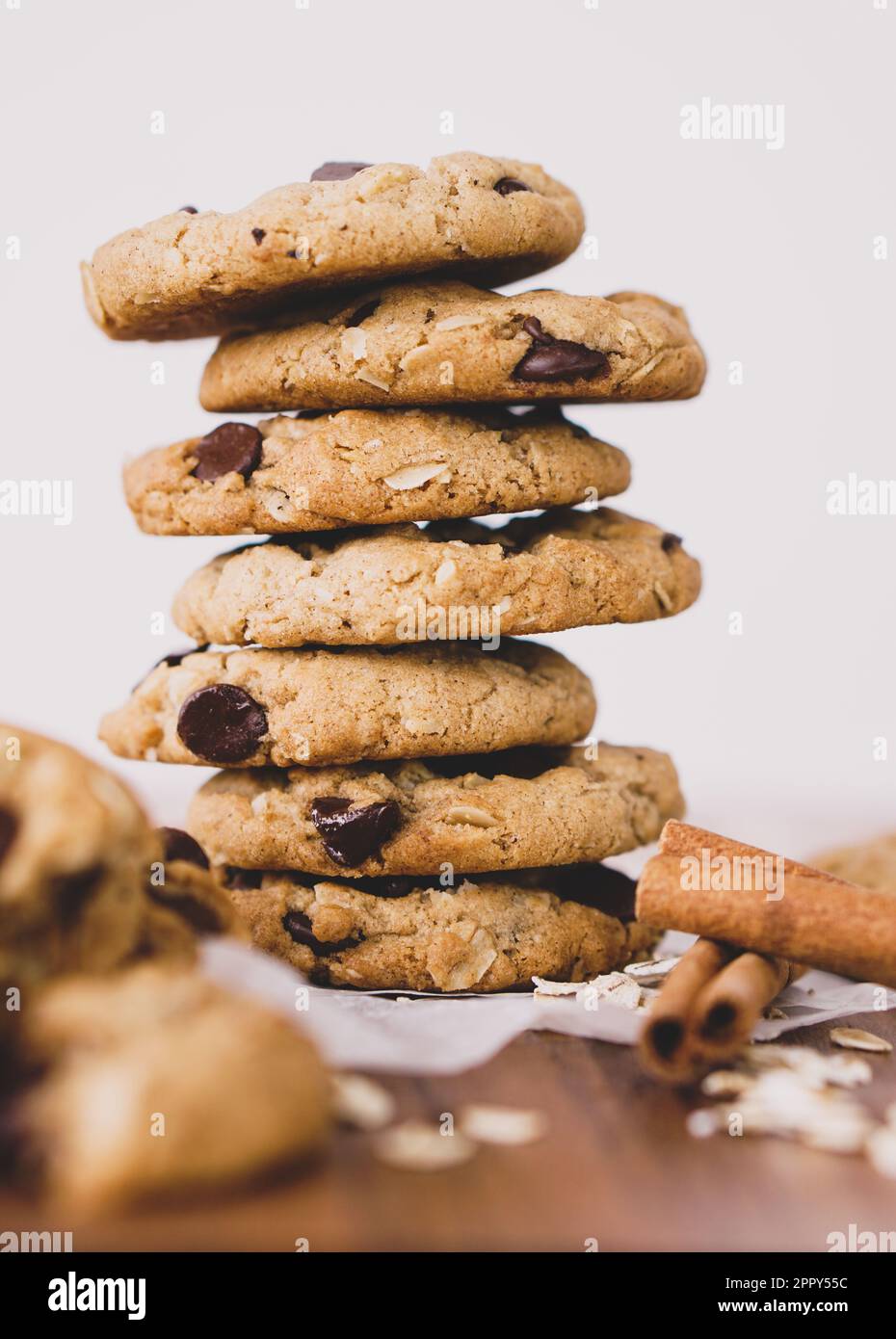 Una pila di biscotti a base di farina d'avena a base di piante vegane con scaglie di cioccolato su un tavolo di legno su uno sfondo bianco. Cannella e avena in primo piano. Foto Stock