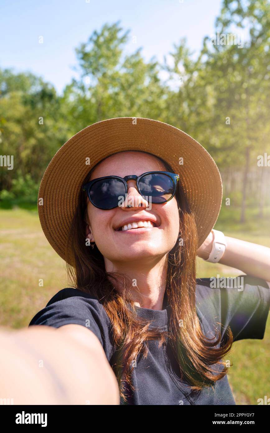Giovane donna con occhiali da sole e cappello di paglia che ride e prende un selfie mentre cammina all'aperto in estate. Foto Stock