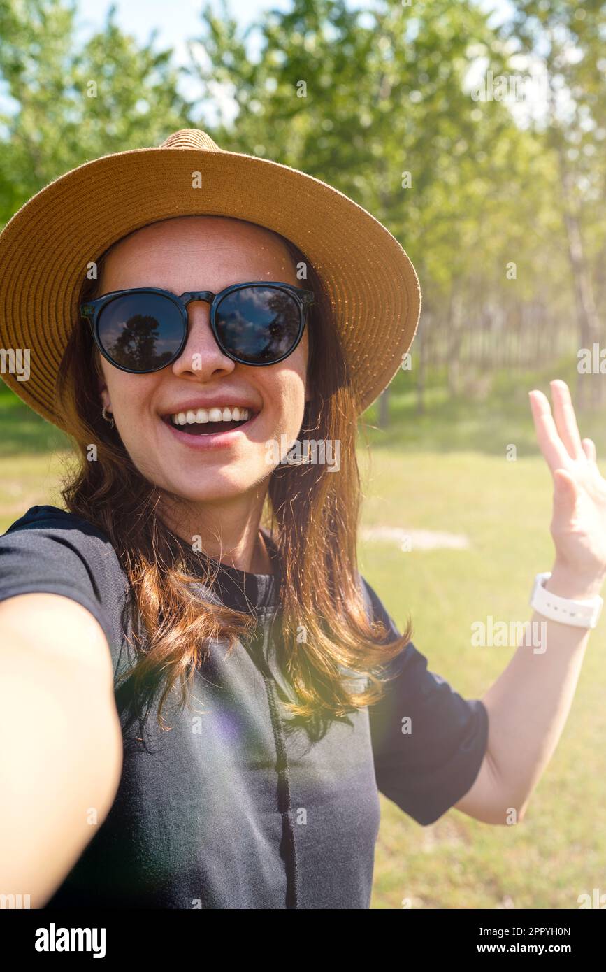 Donna che indossa occhiali da sole e cappello di paglia che agita la mano e che prende un selfie mentre cammina all'aperto. Foto Stock