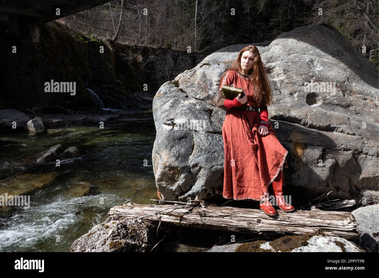 Un modello dai capelli rossi in un vecchio vestito con un libro nelle sue mani si erge su una grande pietra vicino ad un fiume di montagna veloce e turbolento Foto Stock