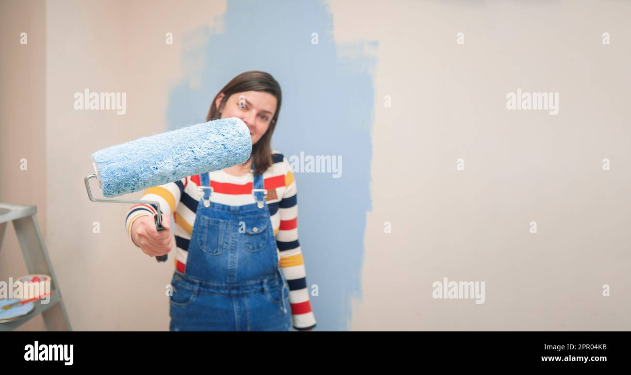 Primo piano di rullo con vernice blu nella mano di una donna in piedi vestita di tute e camicetta a righe, visto dal davanti, sullo sfondo di Foto Stock