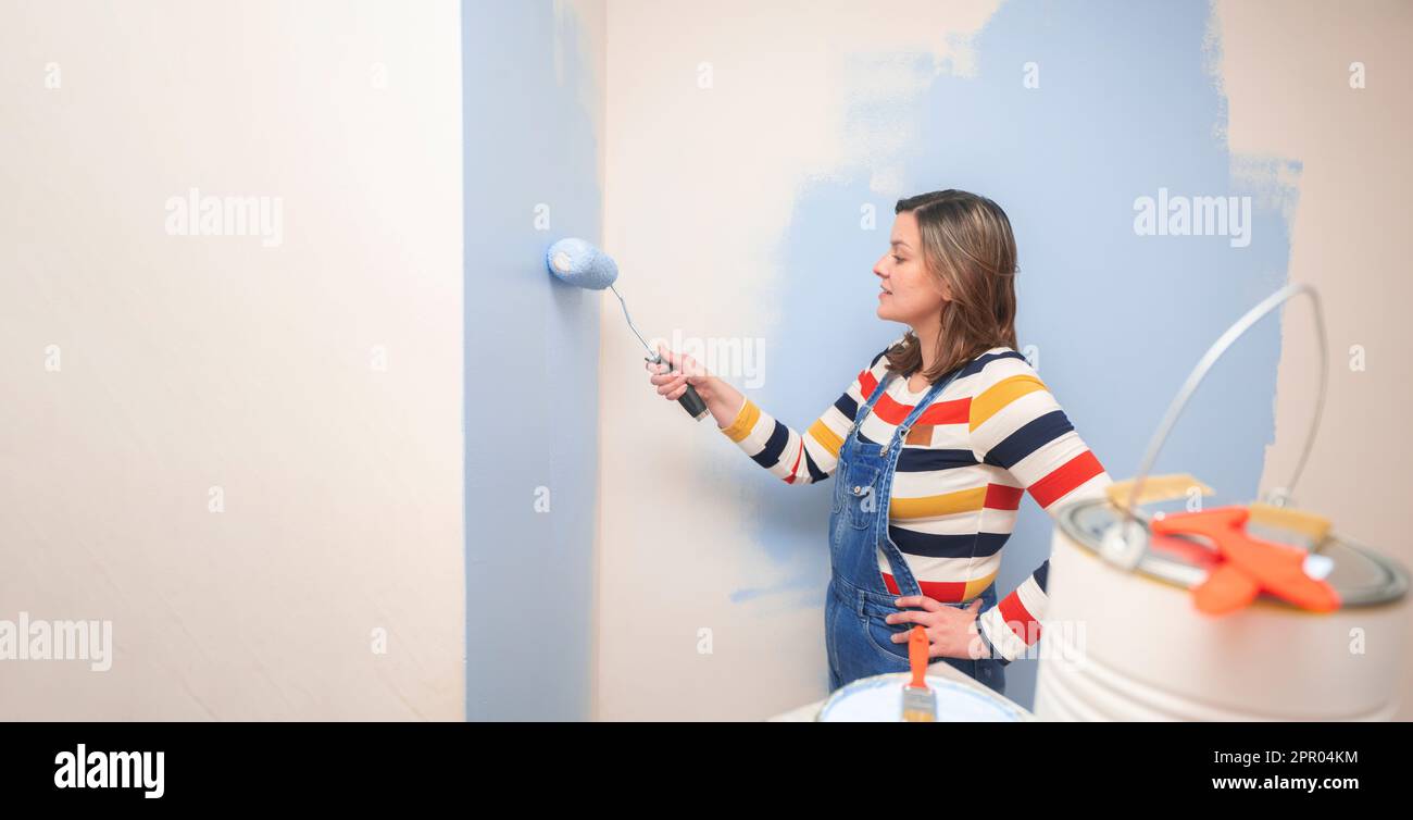 Bella donna in piedi vestita con tute e blusa a righe, vista del profilo, con un rullo in mano dipingendo una parete bianca con vernice blu Foto Stock