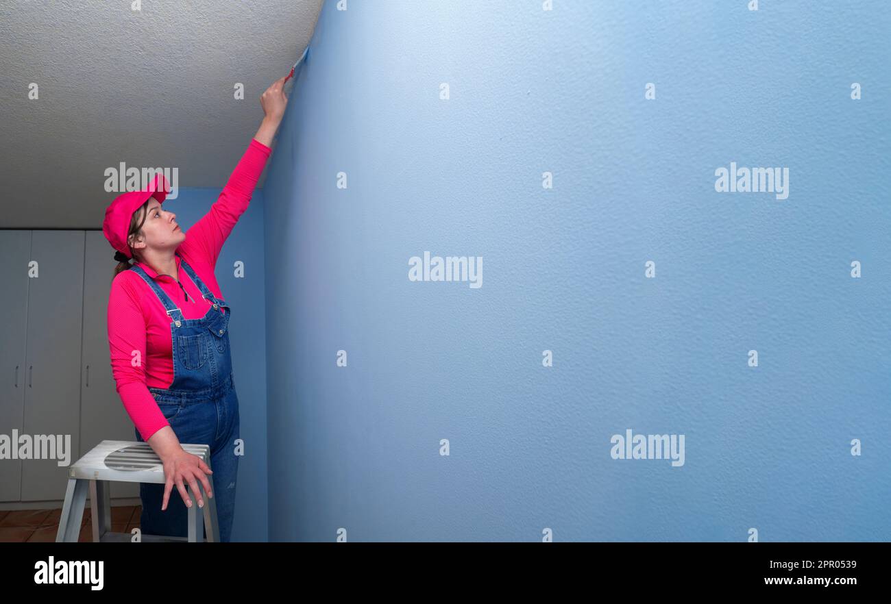 Bella donna vestita di tute e una t-shirt rossa su una scala di metallo, con un pennello in mano che finisce di dipingere un muro blu in una stanza vuota Foto Stock