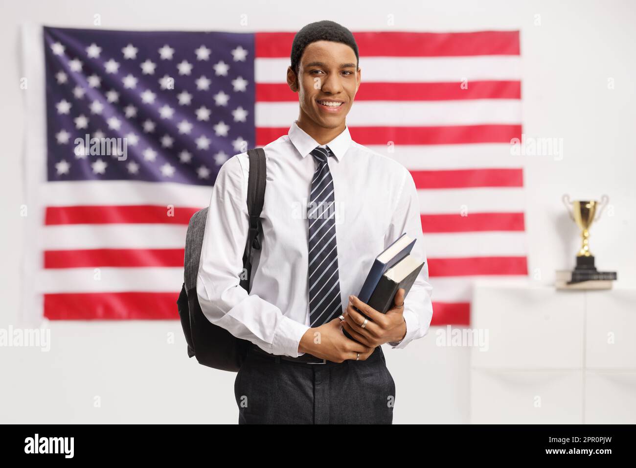 Studente maschio afro-americano in una camicia e cravatta che posa e tiene libri di fronte alla bandiera degli Stati Uniti Foto Stock